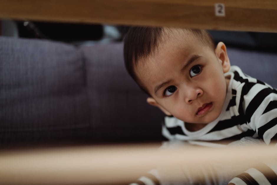 A young child in a striped shirt looks curiously from under a table, indoors.
