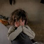 A child in a dimly lit room looking worried, with messy background.