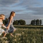 Two happy young sisters running hand in hand through a sunlit meadow.