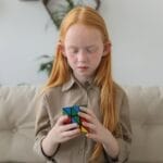 A red-haired girl concentrating on solving a colorful puzzle cube indoors.