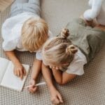 Two young children study together on the floor, concentrating on a notebook in a warm indoor setting.