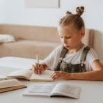 A young girl in a peaceful home setting focused on studying and writing in notebooks.