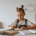 Young girl with braided hair concentrating on her homework at a desk with books, highlighting learning at home.