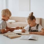 Blonde boy and girl studying together indoors, focusing on writing and learning.