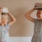 Two smiling children balancing books on their heads indoors, symbolizing fun in learning.