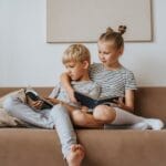 Two siblings sitting on a sofa reading books together indoors.