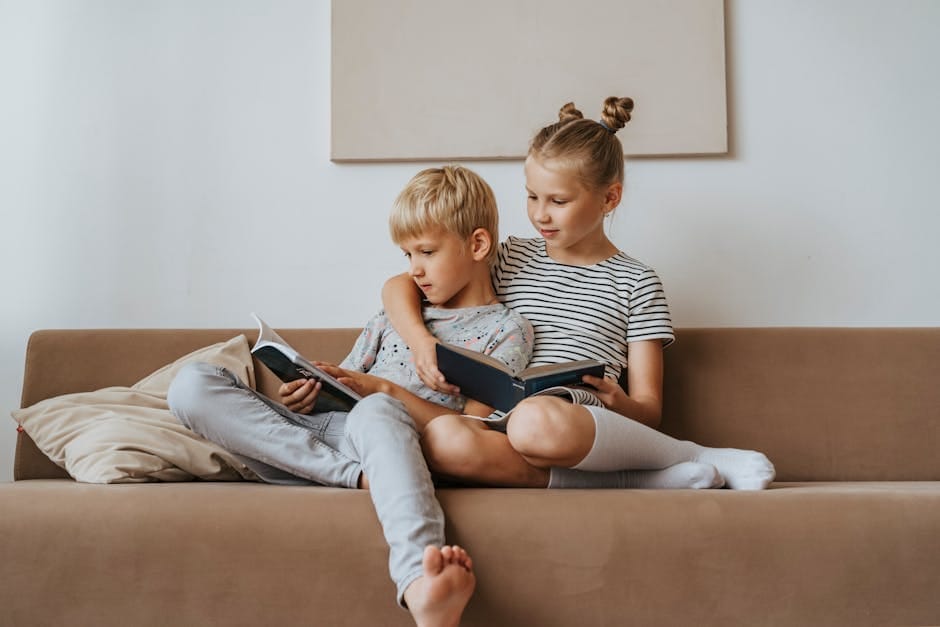 Two siblings sitting on a sofa reading books together indoors.