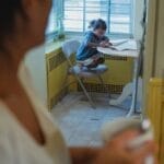 A mother holding a mug watches her child drawing at a table by the window in a cozy home atmosphere.