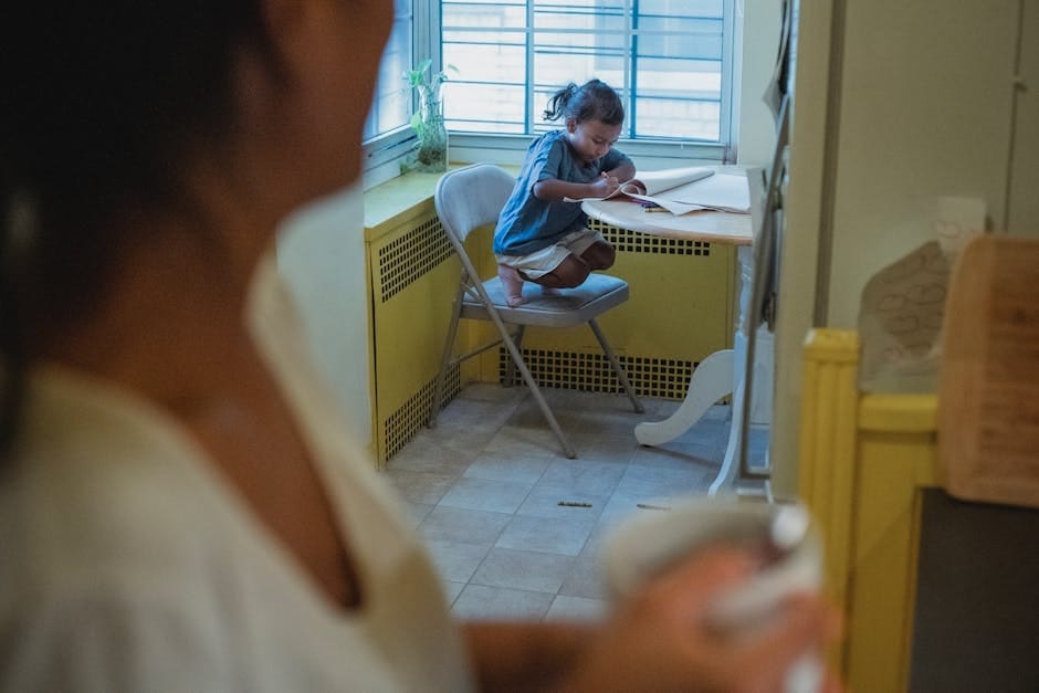 A mother holding a mug watches her child drawing at a table by the window in a cozy home atmosphere.