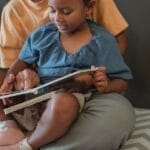 Unrecognizable ethnic mother hugging little girl and reading interesting book while sitting on couch near wall during weekend in apartment