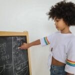 Side view of pensive black girl pointing at mathematical example on blackboard at light room in daytime