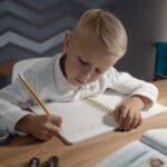 A young boy diligently writing homework in a notebook at a wooden desk indoors.