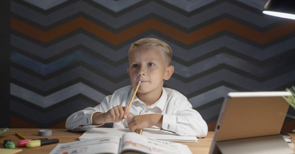 Young boy sitting at desk with open books and tablet, pondering while doing homework under a desk lamp.