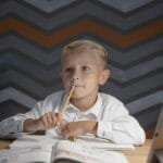 Young boy sitting at desk with open books and tablet, pondering while doing homework under a desk lamp.