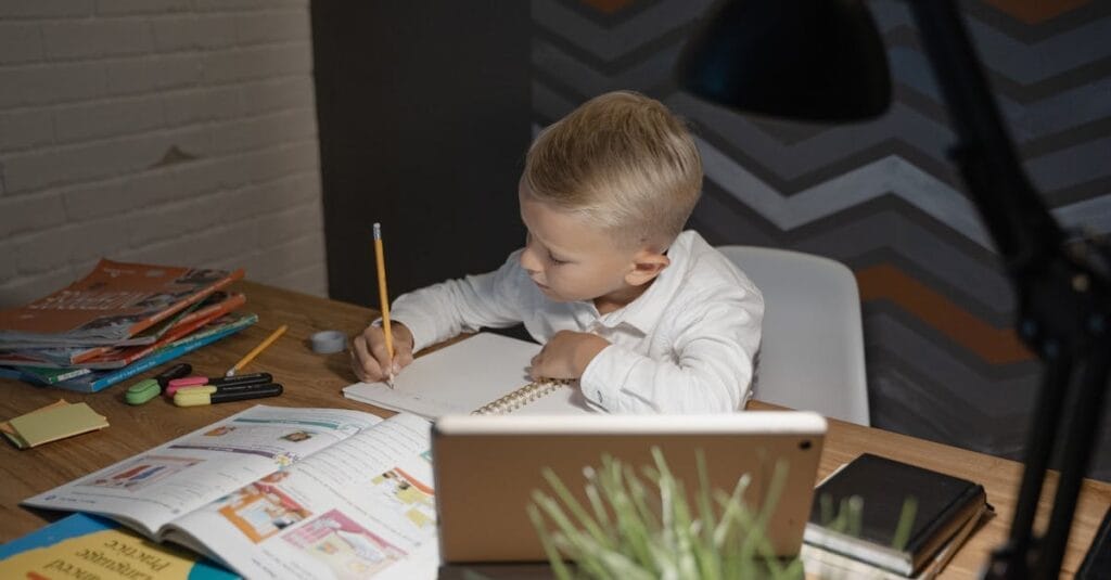 A young boy focuses on homework with books and a tablet at a desk, enhancing his education.