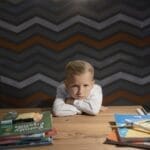 A young boy leaning on a table surrounded by books and coloring materials, looking unhappy with studying.