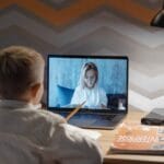 A child participates in an online lesson from home, using a laptop for video conferencing with a teacher.