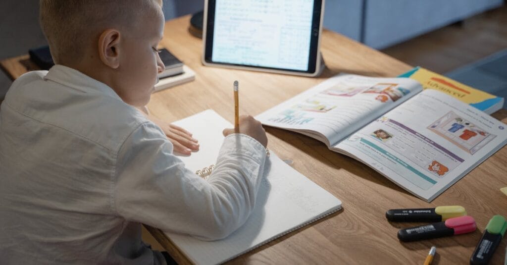 Young boy studying with notebook, pencil, and tablet on a wooden desk, focusing on education.