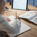 Young boy studying with notebook, pencil, and tablet on a wooden desk, focusing on education.