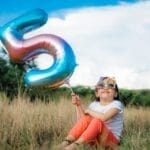 Young girl sitting outdoors holding a colorful number five balloon, wearing fun eyewear, enjoying a sunny day.