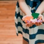 Child displaying colorful paper origami shapes in hands, indoors.