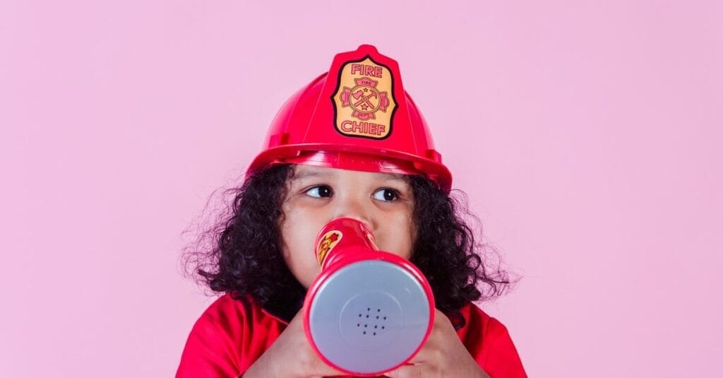 Cute little African American girl wearing bright firefighter costume and helmet and talking in toy loudspeaker while playing in light studio and looking away