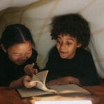 Two children read a book together under a cozy blanket fort, illuminated by soft lighting.