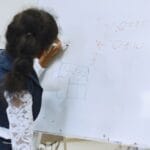 A student writes mathematical equations on a whiteboard in a classroom setting.