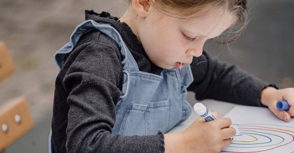 Crop kid sitting on wooden bench at table and holding marker while drawing rainbow