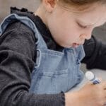 Crop kid sitting on wooden bench at table and holding marker while drawing rainbow