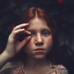 A striking close-up portrait of a young freckled redhead girl holding a pebble to her eye with a mysterious expression.