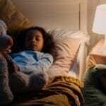 Young girl resting in bed with a teddy bear and toys, illuminated by a bedside lamp.