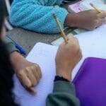 Crop faceless ethnic children sitting at table with pencils and doing homework together