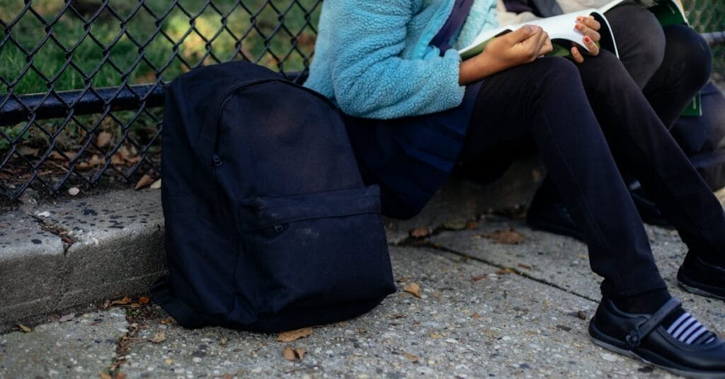 Children sit by a fence reading books, with backpacks on the ground. Autumn setting.