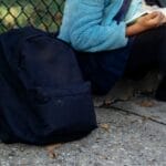 Children sit by a fence reading books, with backpacks on the ground. Autumn setting.