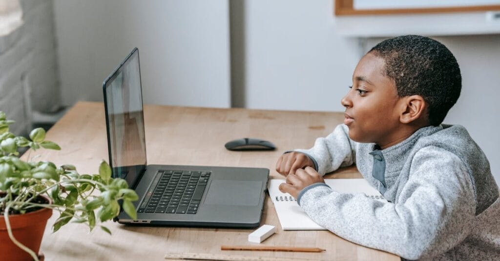 Side view of African American boy watching educational video with netbook while sitting at table with stationery