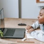 Side view of African American boy watching educational video with netbook while sitting at table with stationery