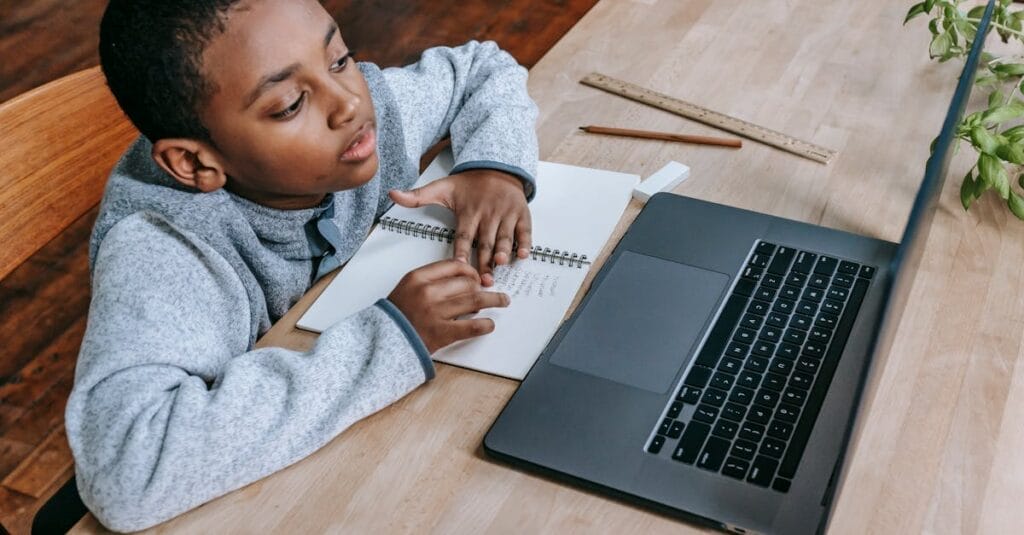 From above of smart cute African American boy studying online with netbook while sitting at table with notebook and pencil