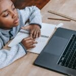 From above of smart cute African American boy studying online with netbook while sitting at table with notebook and pencil