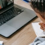 Child engaged in online education using a laptop and taking notes in a notebook.