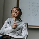 Young African American boy pensively studying in a classroom with math equations on a whiteboard.