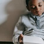 Young boy attentively reading a book while sitting indoors on a wooden floor.