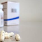 Close-up image of white capsules with medicine bottle and box in the background.