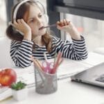 A child wearing headphones doing homework on a laptop indoors. Remote learning setup.