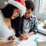 A mother and her son enjoy a festive moment writing Christmas cards by a window.