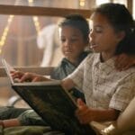 Two siblings enjoying a storybook together, surrounded by cozy lighting indoors.