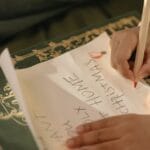 A child writes a handwritten Christmas letter using a red pencil on a piece of paper.