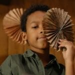 Child holding an artistic paper fan indoors, wooden backdrop.