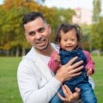 A loving father holds his joyful child with Down syndrome in a park, surrounded by greenery.