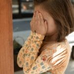 A shy child with brunette hair covers her face while standing outdoors in a playful moment.